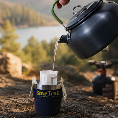 A hand pours hot water from a dark gray kettle into a Star Trail coffee dripper set in a camping environment with a mountain and lake background.
