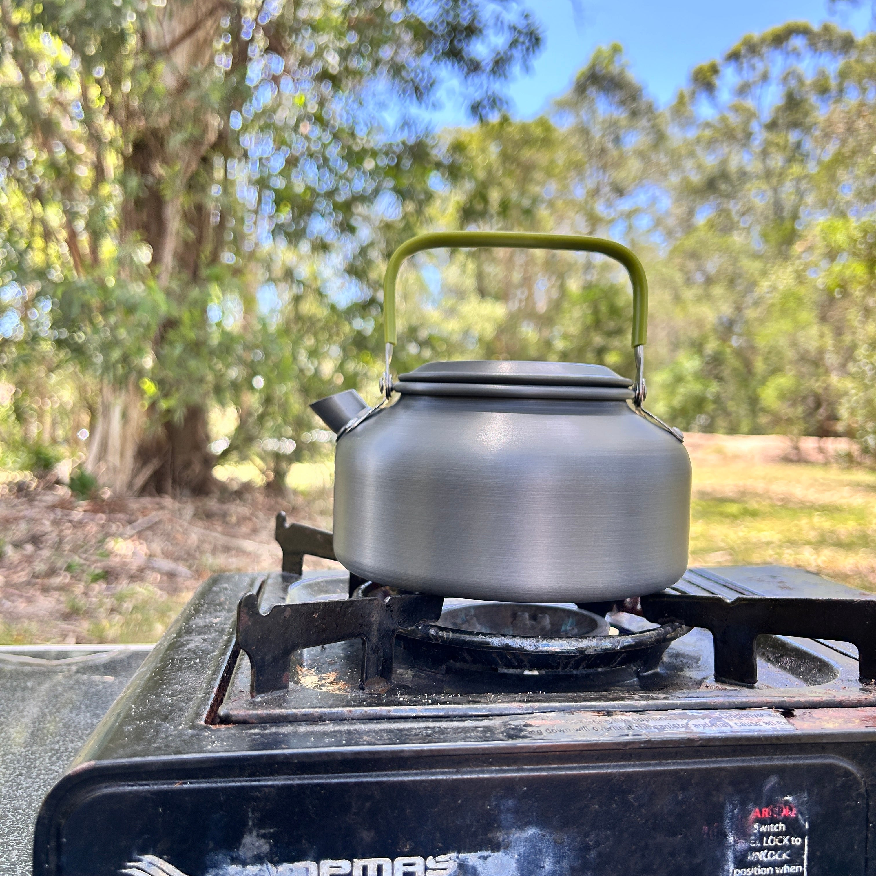 Silver kettle on a camping stove with trees in the background